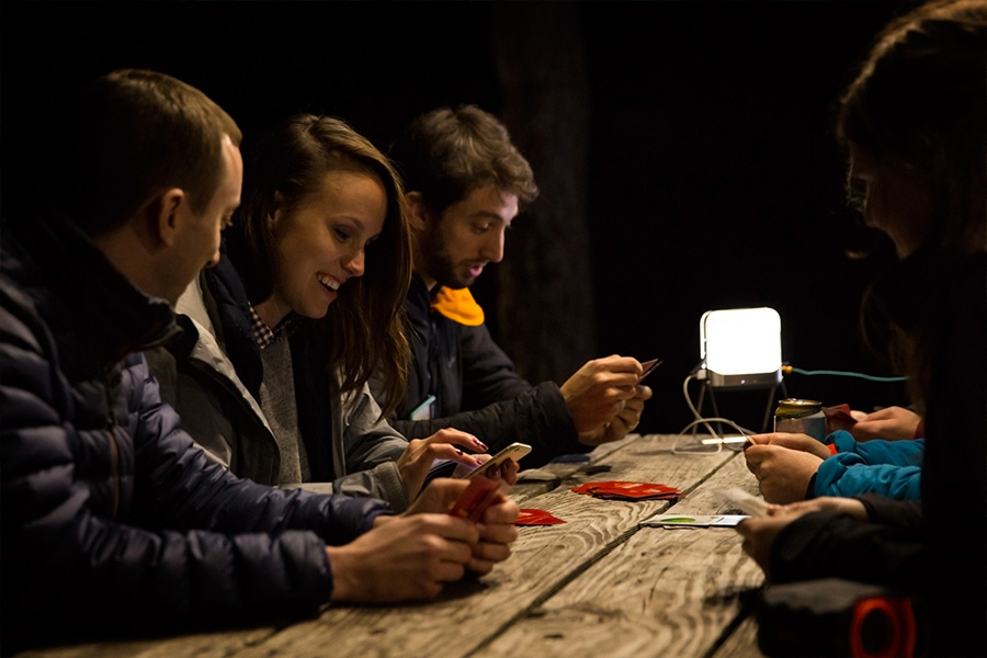 A group of young campers laughing on their phones at a camp table, lit by a lantern.