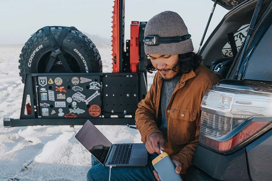 A man in a beanie and jacket is charging his laptop with a power pack, sitting on the back of his 4WD on the beach.