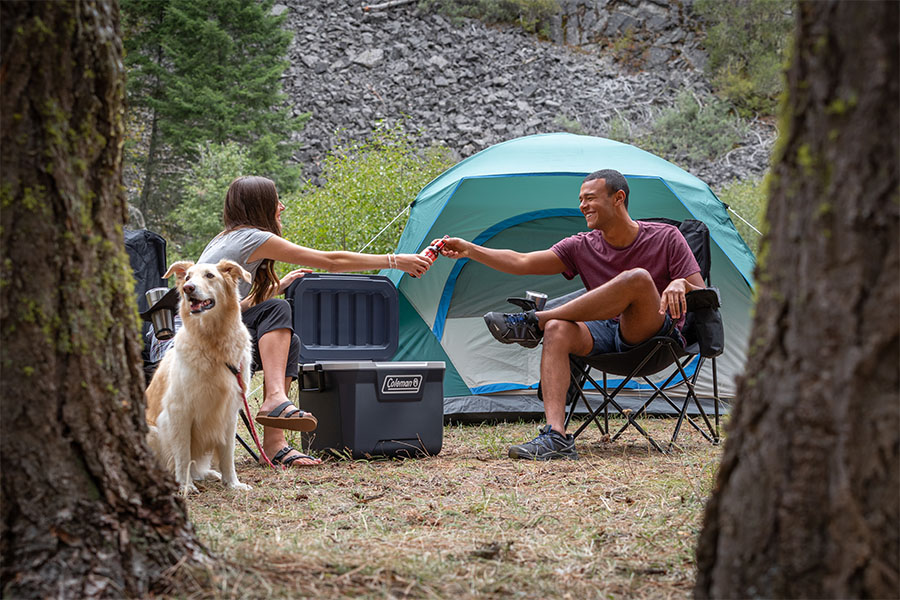 Two campers at a campsite holding a bottle of wine, with a dog sitting beside them and a tent in the background.