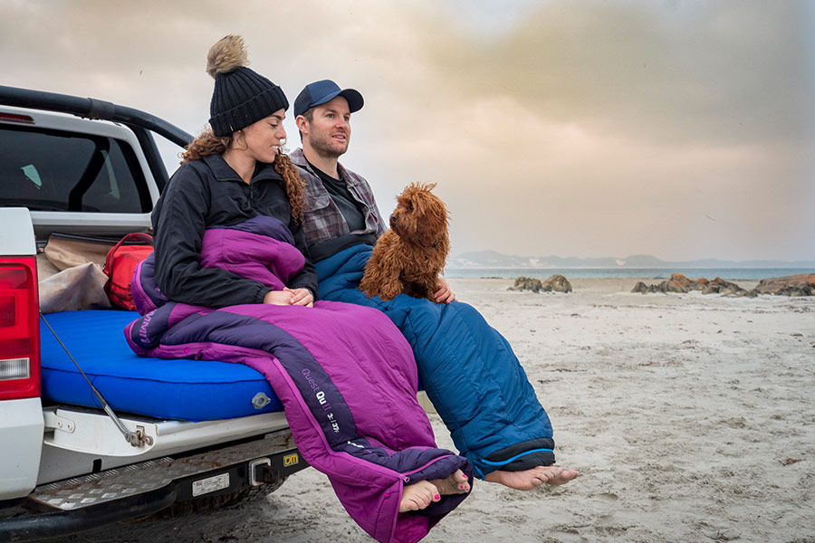 Two campers in sleeping bags, sitting with their dog on the back of their 4WD on a sandy beach.
