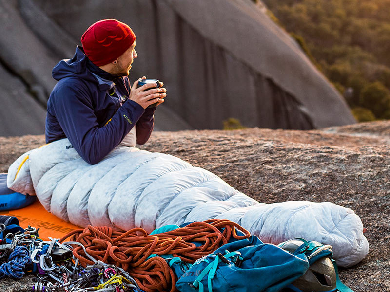 A man in a beanie holding a thermos, snuggled up in a sleeping bag. 