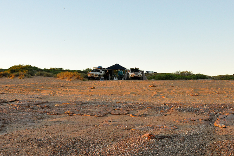 A sandy, pebbly beach in the sunset with two 4WDs and a camp setup in the background.
