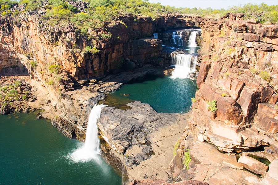 Two waterfalls tipping into two blue pools of water, surrounded by bright red rock and green shrubbery. 