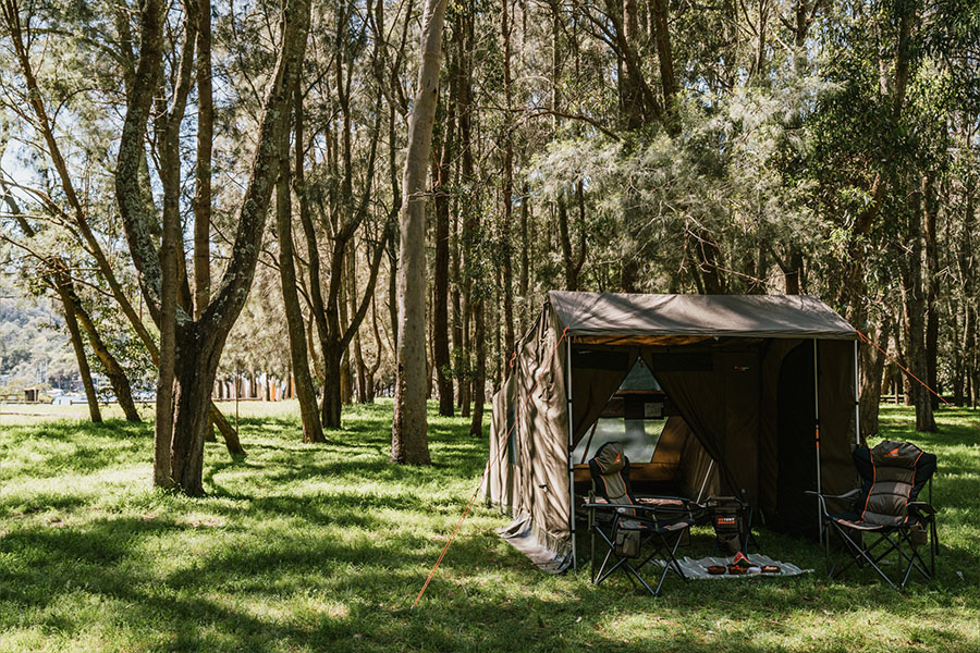 A tent is standing in a forest of trees, the sunlight streaming in, with two camp chairs and picnic set-up out the front. 