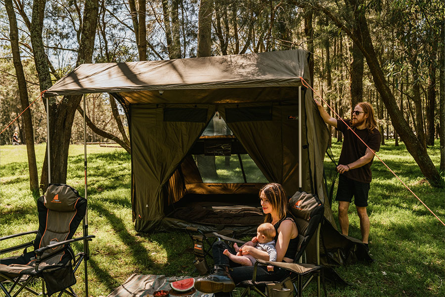A lady sitting with a baby on her lap outside a tent, with watermelon on a picnic rug. A man is pulling back the tent fly. 