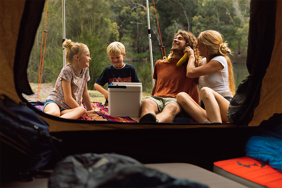 A young family of four sitting on a picnic rug with a cooler, outside a tent. 