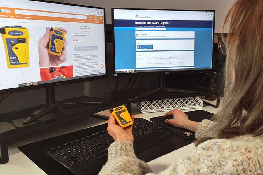 Image shows 2 computer monitors with a woman sitting looking at the screens. She's holding a PLB device and filling out the online registration form.