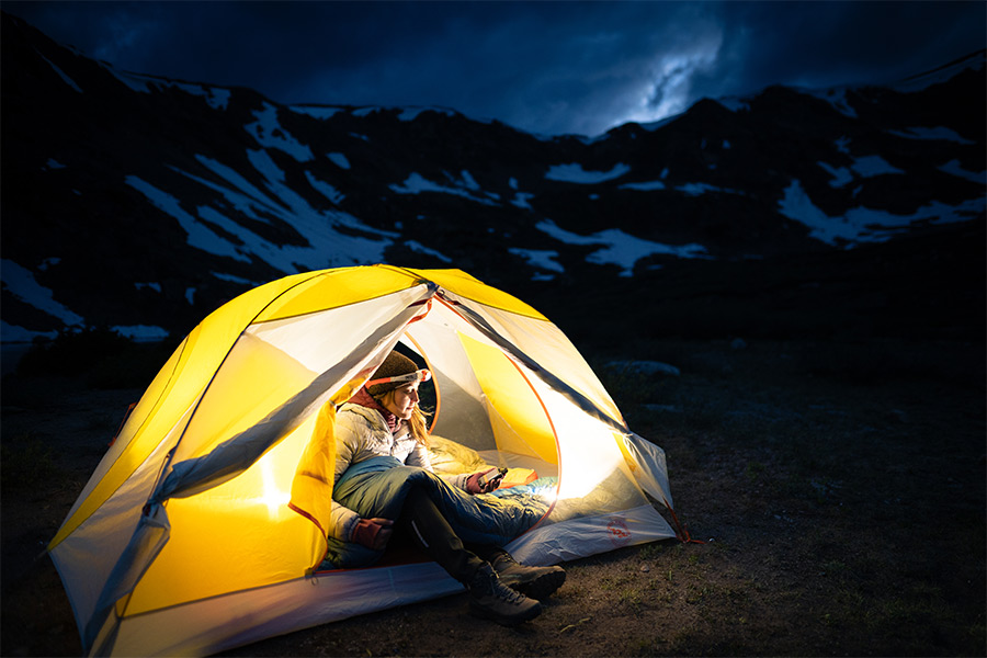 A night time scene in the alps with a female hiker inside her hiking tent. The tent is illuminated by torch light and is glowing yellow.