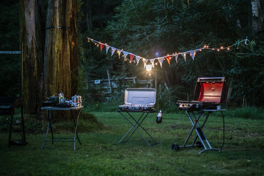 A twilight party scene with green grass, trees and foliage. There's a camp table set up and cookers, plus colourful bunting a lantern and fairy lights.