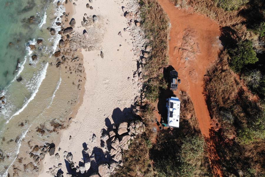 A bird's-eye-view of the ocean, the sand, rocks, and red dirt tracks, with a white caravan and 4WD.