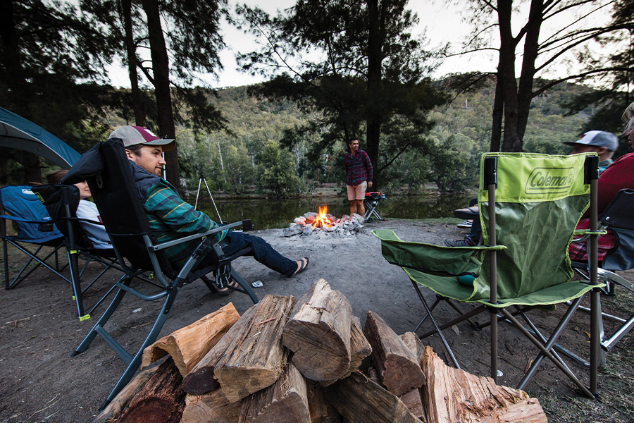 Campers sitting around a campfire with a pile of fire wood nearby.