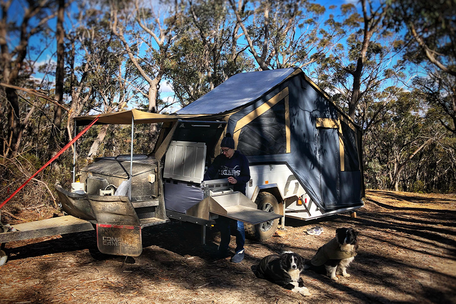 A camper trailer set up with a man and two dogs sitting nearby.