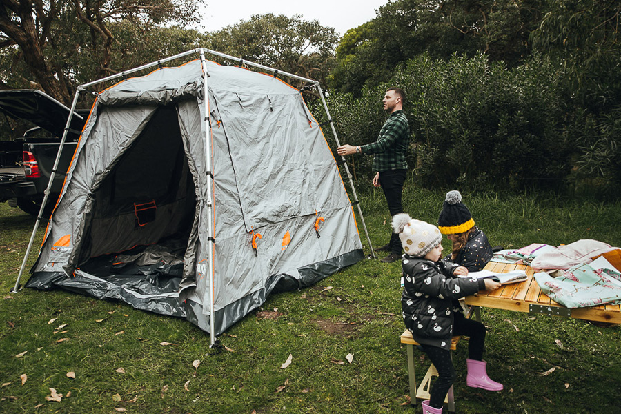 A man setting up a family tent, with two children sitting at a camp table nearby rugged up in beanies and boots.