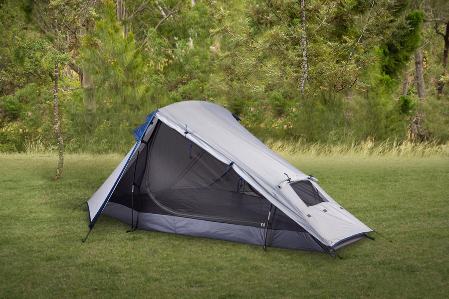 A 2 person hiking tent pitched on green grass with trees behind.