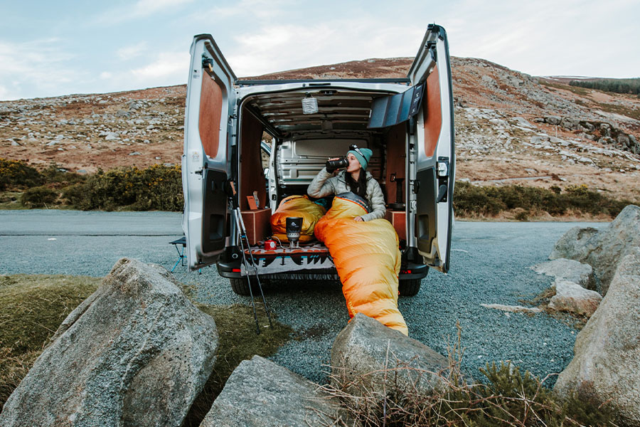 A camper in a sleeping bag, sitting on the back of her van drinking from a Thermos. 