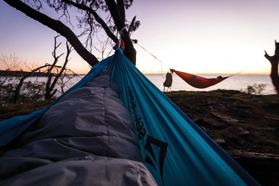 A sleeping bag in a hammock, with another camper in a hammock nearby. It is sunset.