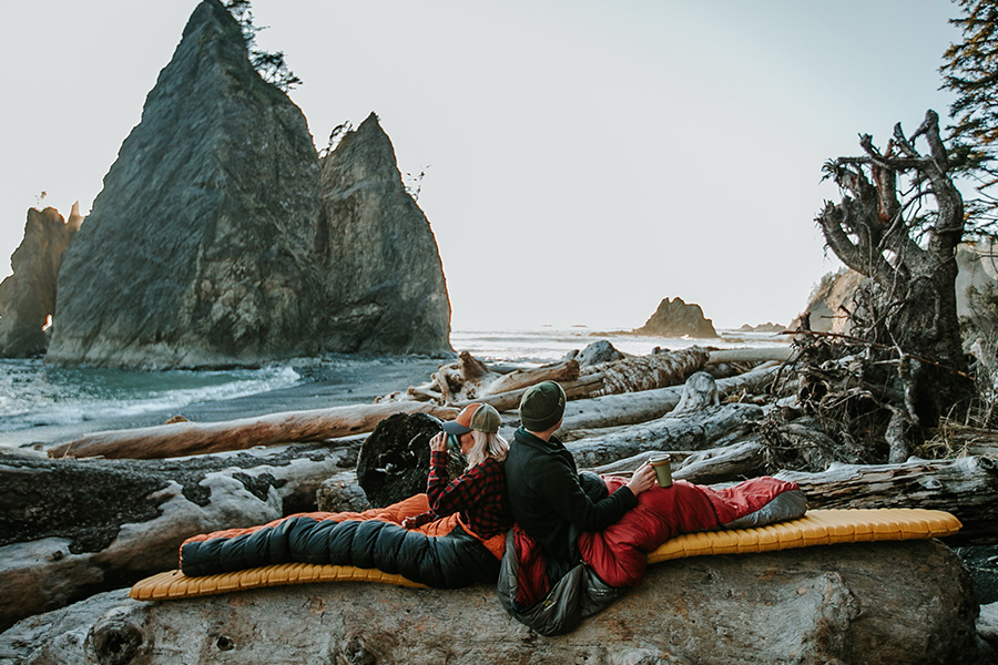 Two campers are sitting back to back in sleeping bags, sipping on hot beverages. 