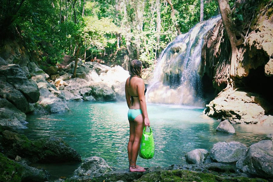 A woman in bathers standing in front of a waterfall with a Scrubba Washbag.