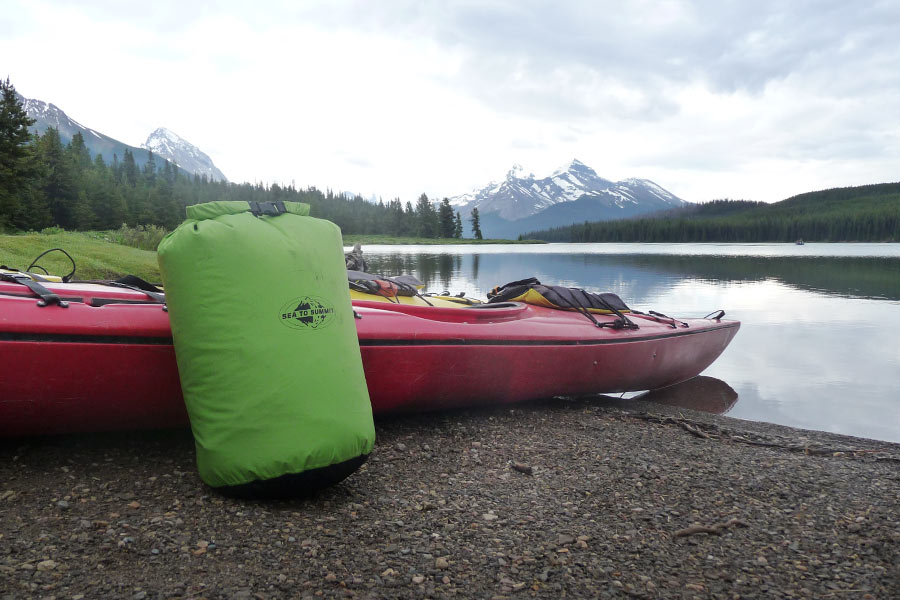 A full dry bag sits next to a kayak at the edge of a river