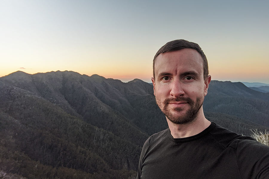 A man smiles at the camera while taking a selfie in front of a mountain peak at sunset