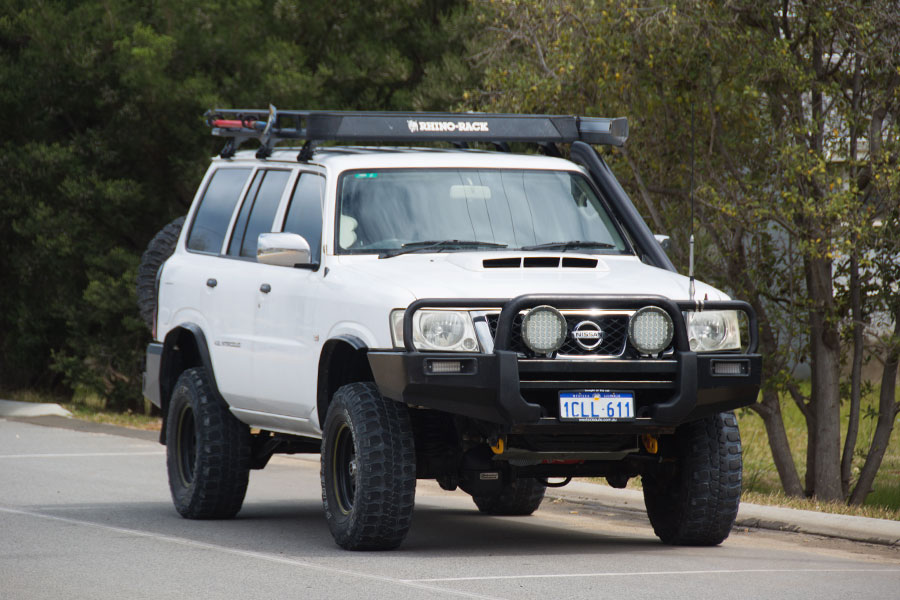 A white Nissan Patrol with LED spotlights is parked by the side of a road.