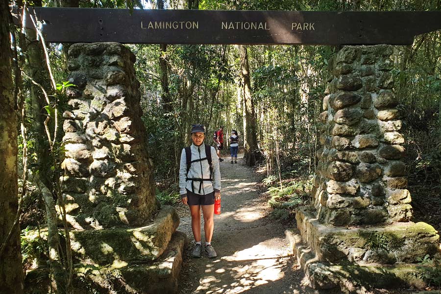 A woman stands beneath a wooden sign fastened atop rock pillars, that reads Lamington National Park