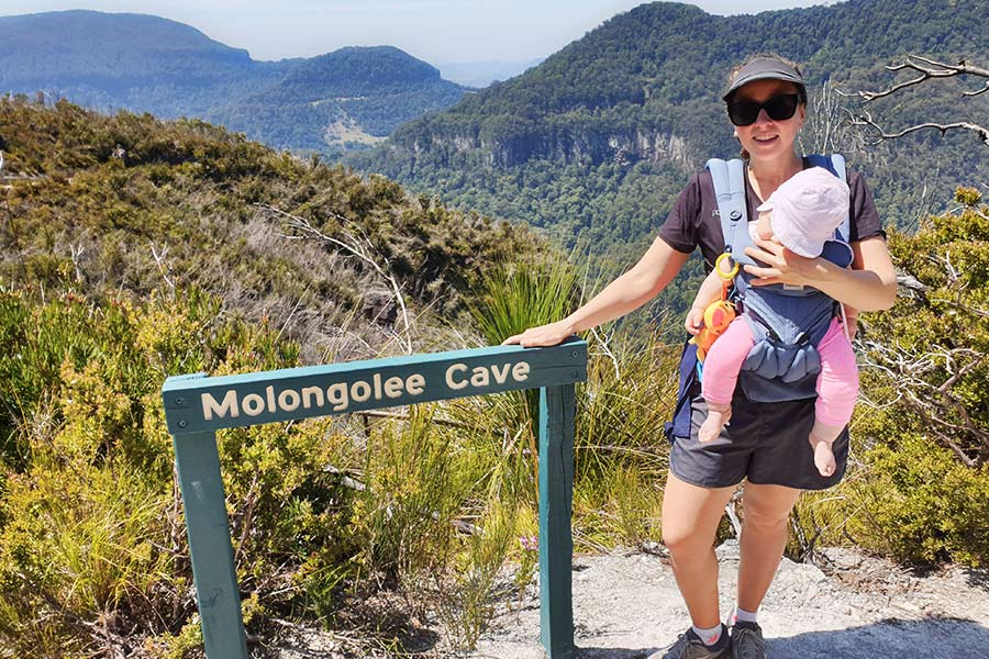 A woman with a baby in a carrier stands on a ridge next to a sign that reads Molongolee Cave