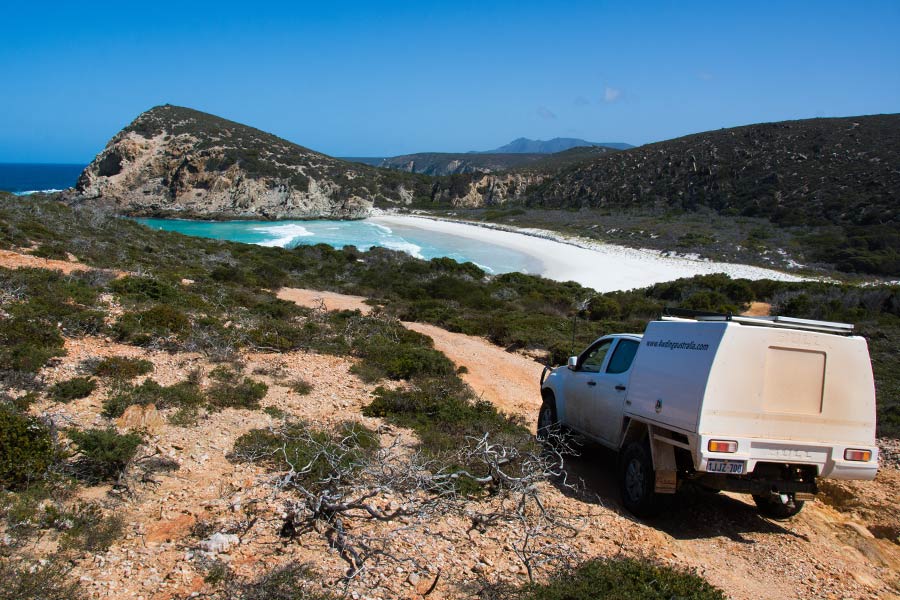 A 4WD drives along a dirt road near a clifftop, with views of an ocean bay below.