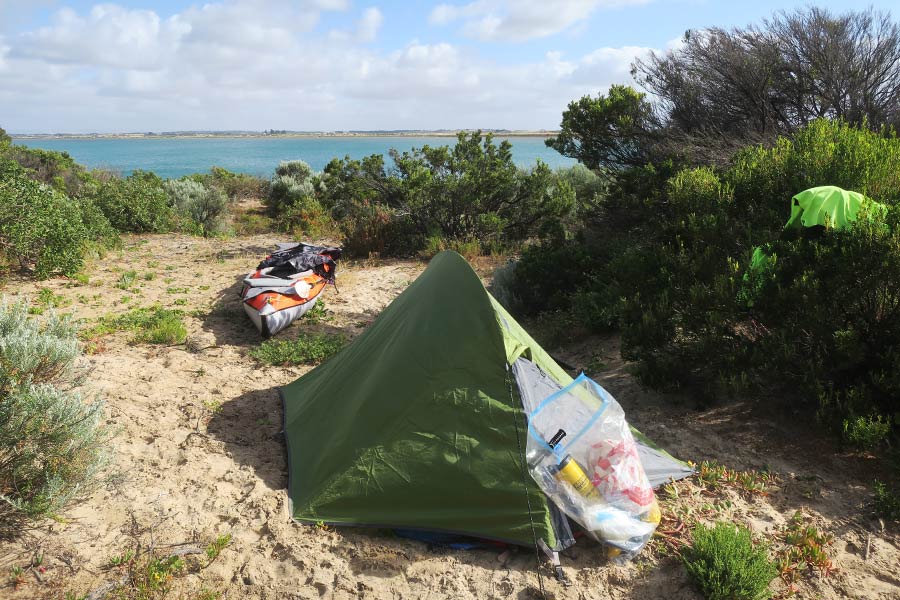 A waterproof bag rests on a tent, set up on sand dunes near a kayak with the lagoon in the distance.