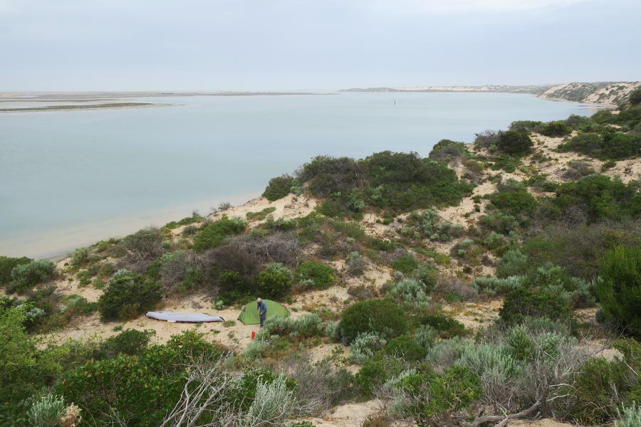 A man stands near a hiking tent and kayak, amongst the sand dunes near a lagoon.