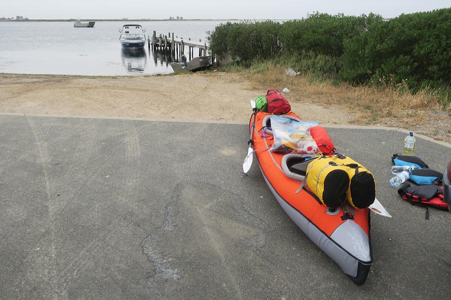 An inflatable kayak, partly packed with gear, sits on bitumen near the water.