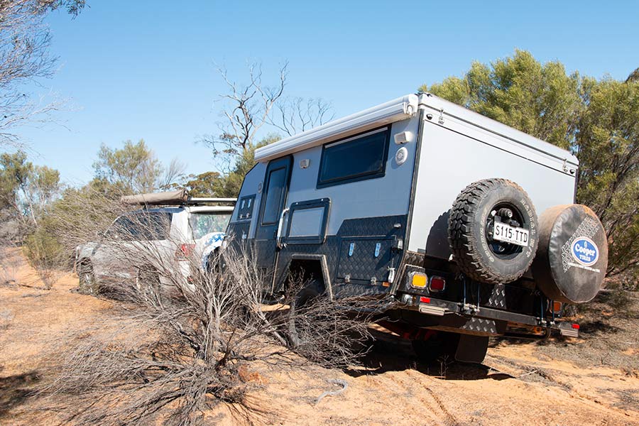 An off-road caravan is towed between trees over remote terrain.