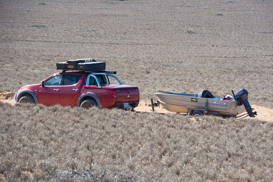 A ute with full roof racks tows a small boat along a dirt road.