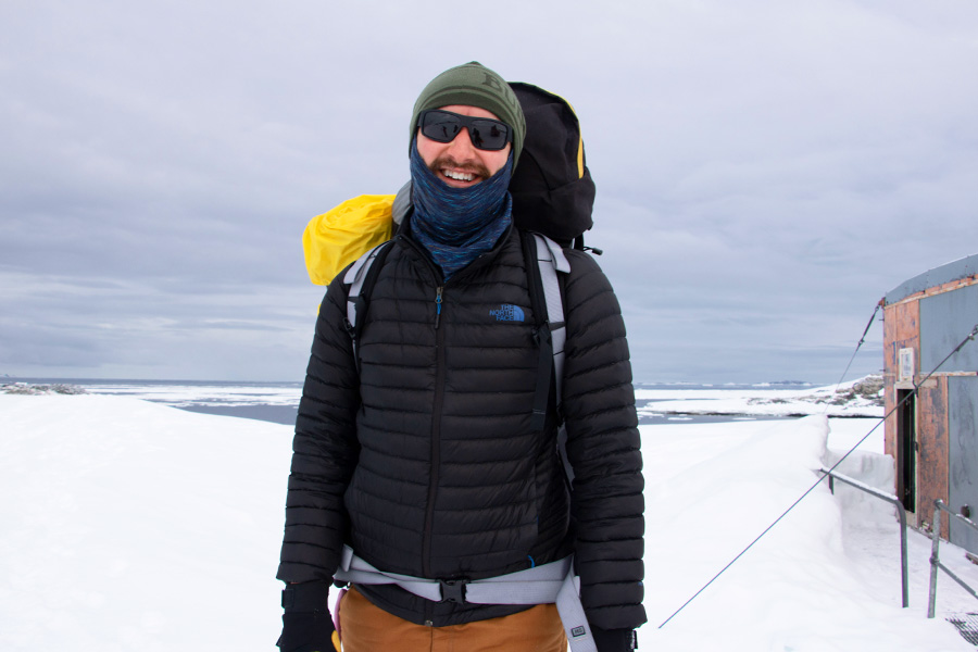 A man standing on snow in Antarctica wearing a padded jacket, neck buff, beanie and large backpack.