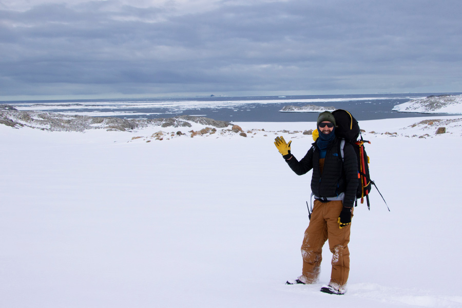 Man in snow gear and carrying a large backpack, waving to the camera with a yellow gloved hand. He is standing on the white snowy ice in Antarctica with grey skies and Arctic waters behind him.