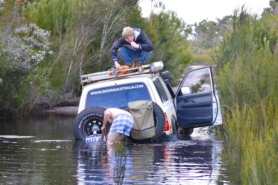 A 4WD vehicle stuck in a deep water crossing with bush shrubbery around. There's a man crouching on the vehicles roof and another man in the water attaching a snatch strap for recovery.