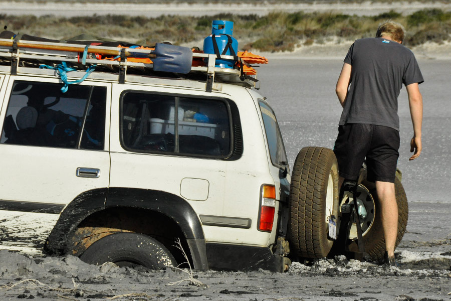 A white 4WD sunk up to the wheel hubs in a salt lake. A man is standing with his back to the camera, looking down at the muddy situation.