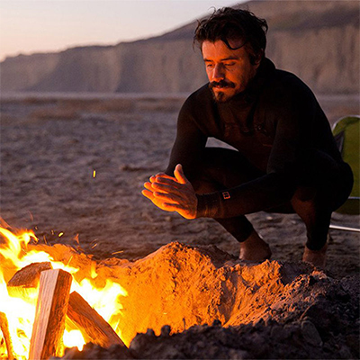 A man squatting beside a campfire on the beach. He's warming his hand over the flames and there are cliffs in the background.