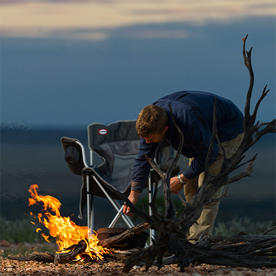 A man leaning over a campfire, stoking it with a large dead branch. It's early evening in the Outback with a folding camp chair nearby.