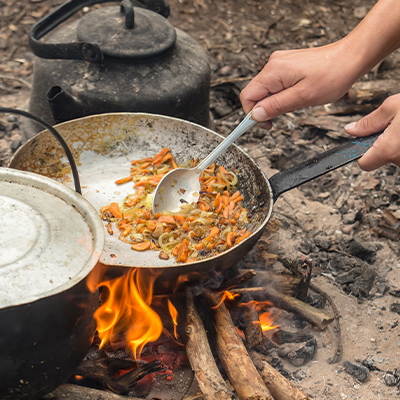Hands holding a frypan over the flames of a campfire and cooking food. There's a lidded pot in the foreground and a blackened kettle in the background.