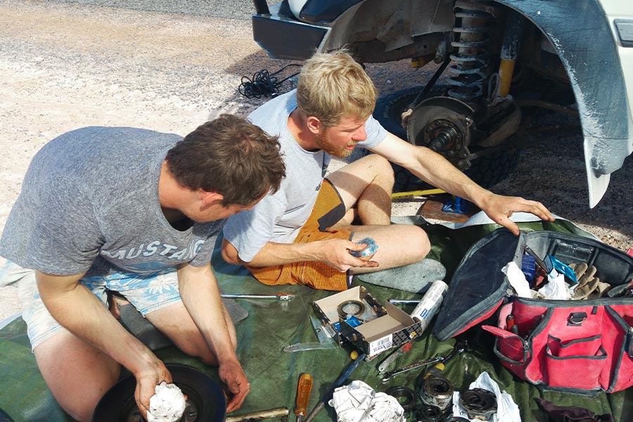 Two men sit beside the wheel hub of a 4x4. They have a drop sheet with tools spread out and the wheel has been removed.
