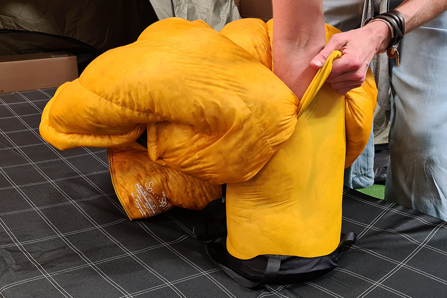 An arm stuffs a yellow sleeping bag into a matching compression sack.