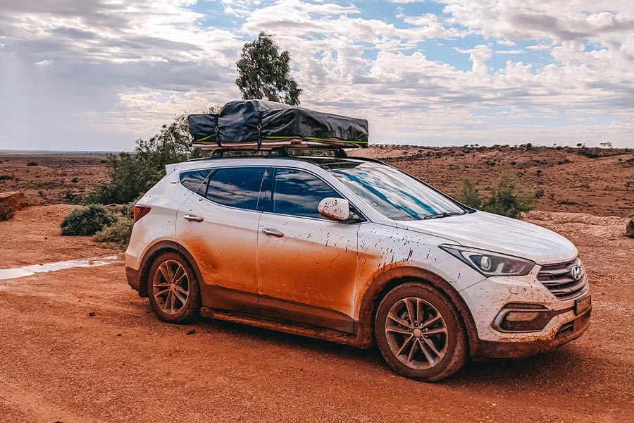 A white sedan with a roof pack is parked in the outback and splattered with red dirt and mud.
