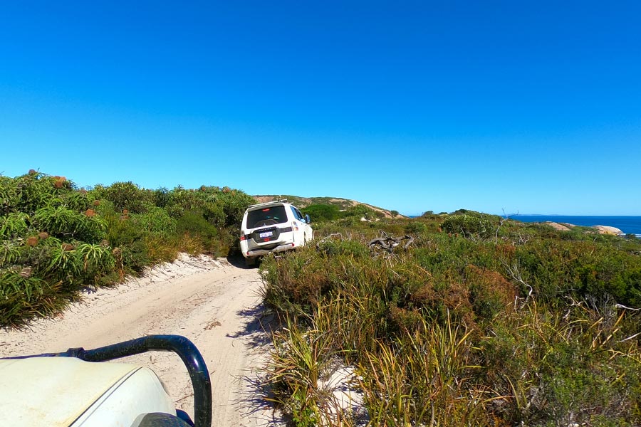 One vehicle follows another at a safe distance along a coastal 4WD track. There's green shrub either side, a bit of ocean to the right and clear blue sky above.