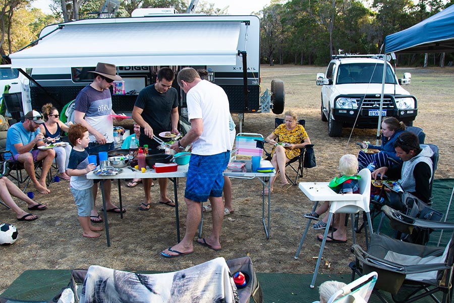 A group of campers sharing a communal breakfast. There's a central table and chairs scattered about, plus a car and caravan behind.