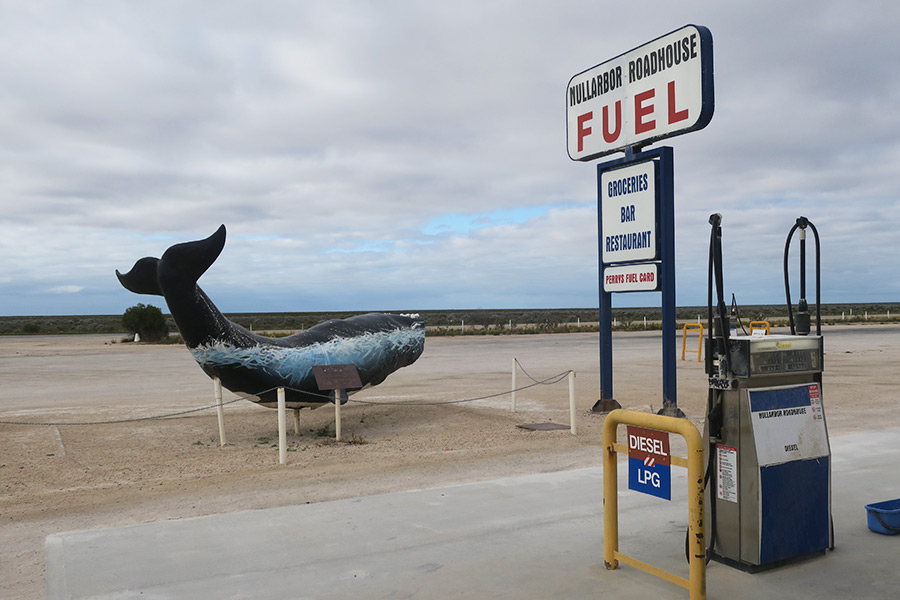 Signage and bowser at the Nullarbor Roadhouse with sculpture of a whale out the front. The Nullarbor Plain stretches off to the horizon.