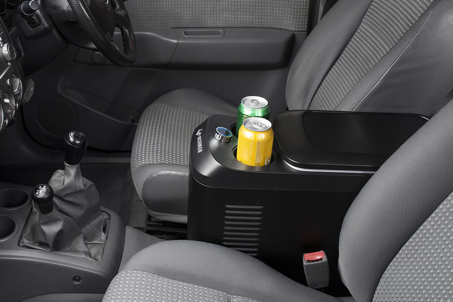 A Bushman car fridge positioned between the 2 front seats of a vehicle. There are 2 cans of drink in the built-in cup holders.