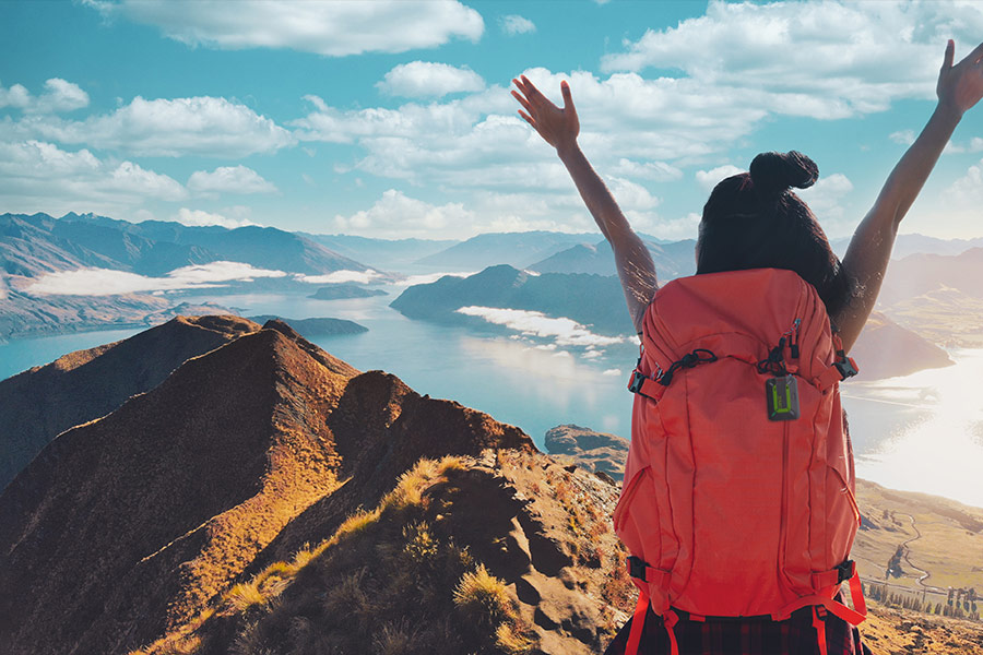A female hiker celebrates reaching the summit of a mountain with her arms out stretched in the air above her head. The view shows her towering over surrounding mountains, and water inlet and even scattered clouds. There are more clouds above and she's wearing a red backpack with a ZOLEO messenger device attached.