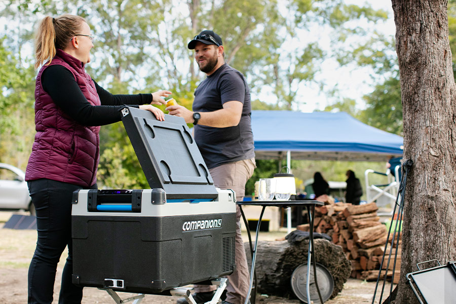 A Companion Lithium 60L Dual Zone fridge set up on a stand at a campsite. A woman in a purple vest is passing a man a can of drink from an open fridge.