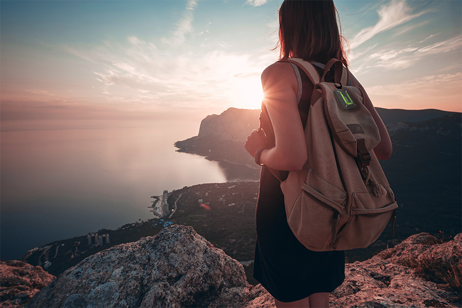 A female hiker stands on the summit of a coastal mountain looking out to sea. There's a beautiful sunrise casting a hazy glow over the water and surrounding landscape, and the woman is wearing a brown canvas backpack with a ZOLEO messenger device attached to the top.